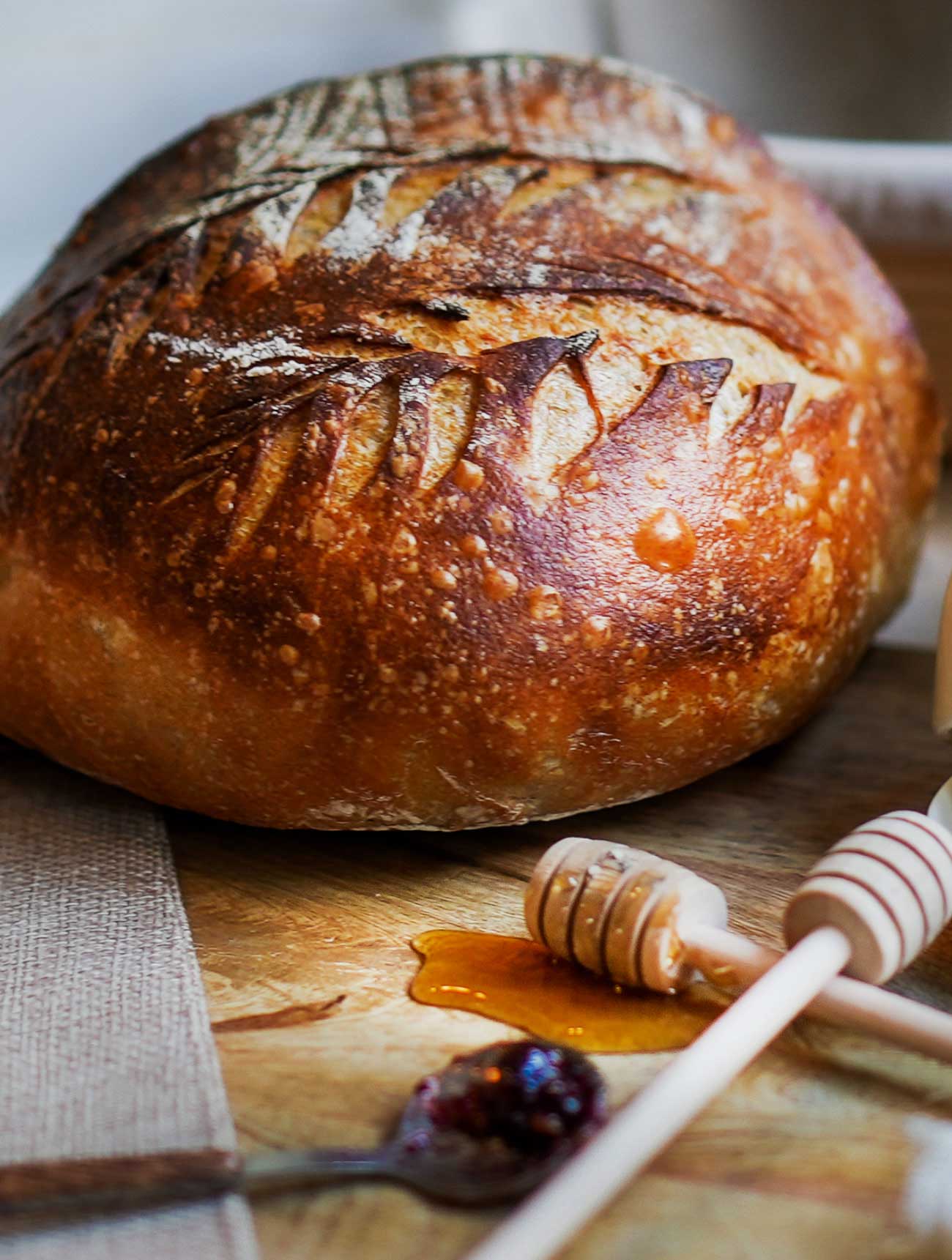 Crusty fail-proof sourdough bread loaf on rustic wooden board with honey swizzle stick and spoonful of strawberry jam