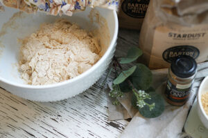 Overhead view of shaggy sourdough dough in a bowl with Stardust flour and a cinnamon bottle on a rustic white board