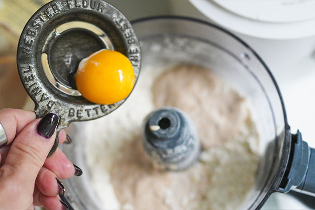 Sourdough beef pot pie in white cocottes flaky crust comfort a close up of a womans hand using an egg separator to add a yolk into a food processor bowl filled with flour butter and sourdough starter The yolk is centered in focus capturing a warm homemade moment in the pie crust making process Sourdough afterglow Woman’s hand holding an egg separator with yolk above food processor, ready to be added to sourdough pie crust ingredients