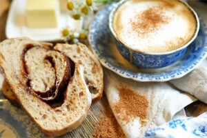 Overhead view of a cinnamon swirl sourdough slice with butter in the background, latte to the right, and sprinkled brown sugar mix nearby