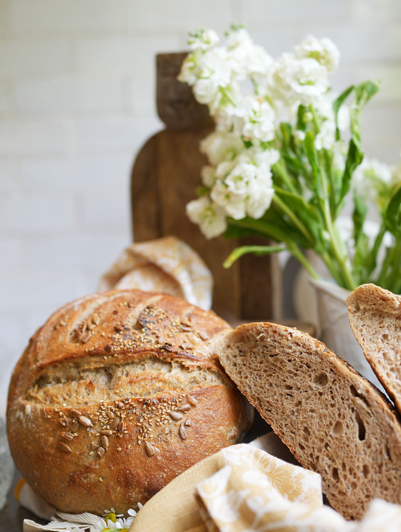 Crispy seeded sourdough with two crumb halves to the right and flowers in the background