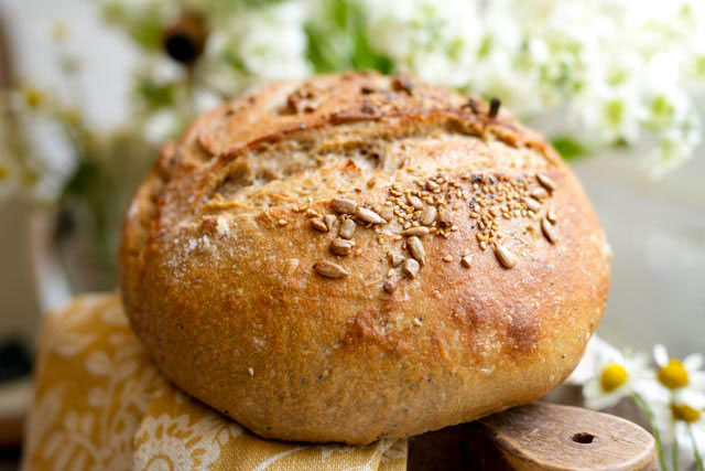 Seeded sourdough bread with crunchy crust and light wheat crumb a beautifully light and crusty sourdough loaf rests on a rustic wooden board with flowers softly blurred in the background The crust is golden and textured hinting at a soft airy crumb inside A warm and inviting image of everyday artisan baking Sourdough afterglow Light crusty sourdough loaf on board with flours in the background