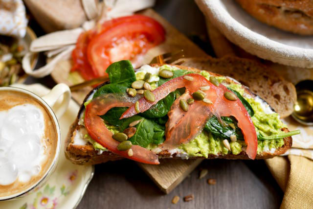 Seeded sourdough bread with crunchy crust and light wheat crumb a slice of seeded sourdough piled with messy smashed avocado and thinly sliced tomato served beside a warm creamy latte The golden crust peeks through the layers and the scene captures the cozy chaos of real breakfast fresh flavorful and perfectly unfiltered Sourdough afterglow Messy avocado toast with tomato slices on seeded sourdough and a latte nearby