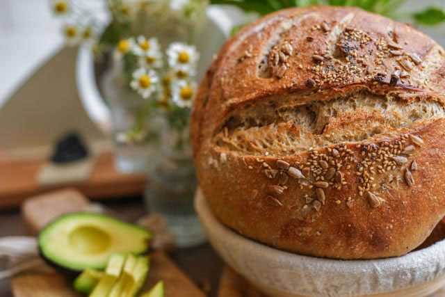 Seeded sourdough bread with crunchy crust and light wheat crumb a sliced seeded sourdough loaf is topped with fresh avocado on a wooden board In the softly blurred background white daisies add a bright natural touch The golden crust and creamy green slices create a fresh nourishing scene simple beautiful and full of flavor Sourdough afterglow Seeded sourdough loaf with sliced avocado on a wooden board and daisies in the background