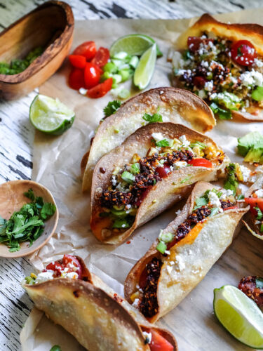 Six crispy chicken tacos on parchment paper with bowls of cilantro, green onion, tomato, and fresh limes ready to serve