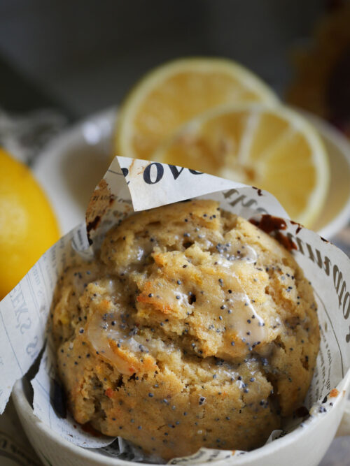 Lemon poppy seed sourdough discard muffin with frosting drizzle in a china cup and fresh lemons in the background