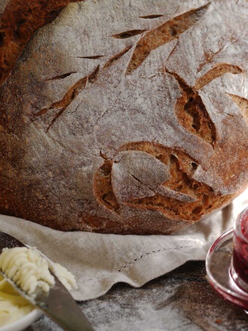 Rustic light whole wheat sourdough bread loaf with floured crust and scoring, served with butter and jam.