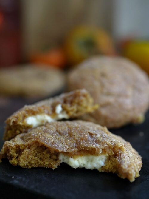 Sourdough pumpkin cheesecake cookie broken open on a dark surface showing creamy cheesecake filling and cinnamon sugar sparkle.